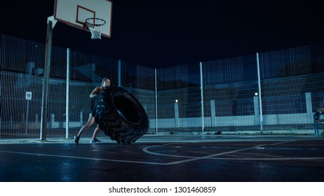 Beautiful Energetic Fitness Girl Is Doing Exercises In A Fenced Outdoor Basketball Court. She's Flipping A Big Heavy Tire In A Foggy Night After Rain In A Residential Neighborhood Area.