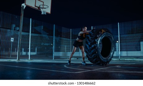 Beautiful Energetic Fitness Girl Is Doing Exercises In A Fenced Outdoor Basketball Court. She's Flipping A Big Heavy Tire In A Foggy Night After Rain In A Residential Neighborhood Area.