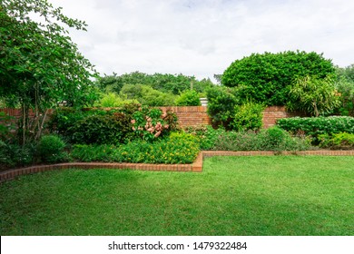 Beautiful English cottage garden, colorful flowering plant on smooth green grass lawn with orange brick wall and evergreen trees on background, in good care maintenance landscaping of a park 