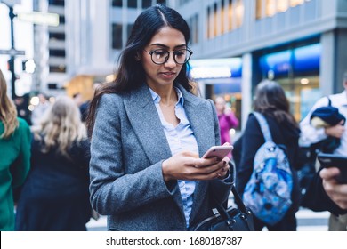 Beautiful Ethnic Business Woman With Black Hair In Glasses And Jacket Browsing In Smartphone While Walking Down Crowded Street In New York City