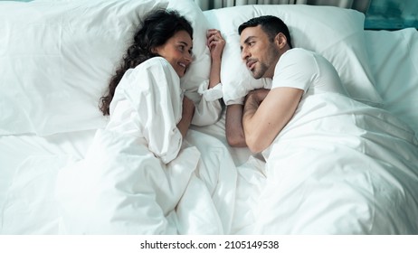 Beautiful Ethnic Couple Laying In Bed Under The Blanket Looking At Each Other And Talking After Waking Up. Celebrating Valentines Day Together In A Fancy Hotel Suite.