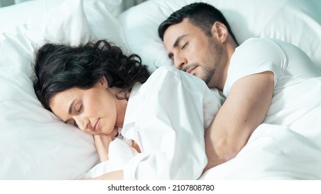 Beautiful Ethnic Couple Laying In Bed Sleeping, Wearing White Pyjamas As They Enjoy  Their Day Off In A Fancy Hotel Room Celebrating Valentines Day. Young Married Couple Enjoying Time Off Together. 