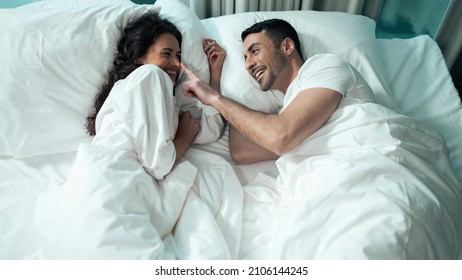 Beautiful Ethnic Couple Laying On The Bed Smiling And Fooling Around. Celebrating Valentines Day Together In A Fancy Hotel Suite. Latino Man Looking In Love With His Latina Girlfriend With Curly Hair.