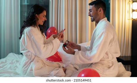 Beautiful Ethnic Couple Sitting On The Bed Of A Fancy Hotel Suite, Smiling In Happiness As The Handsome Latino Man Asks Her To Marry Him. Woman Looking At The Ring In Amazement As The Man Smiles.