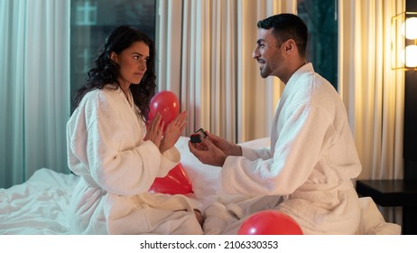 Beautiful Ethnic Couple Sitting On The Bed Of A Fancy Hotel Suite As The Man Wearing A Bathrobe Asks The Female To Marry Him, Holding A Cute Ring Box With An Engagement Ring Inside It.