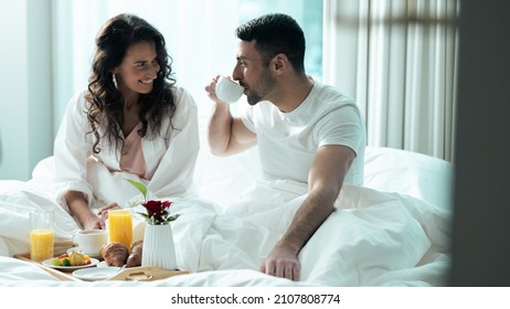 Beautiful Ethnic Couple Sitting On The Bed Eating Breakfast Together And Looking At Each Other In Love. Celebrating Valentines Day Together By Going To A Fancy Hotel Suite.