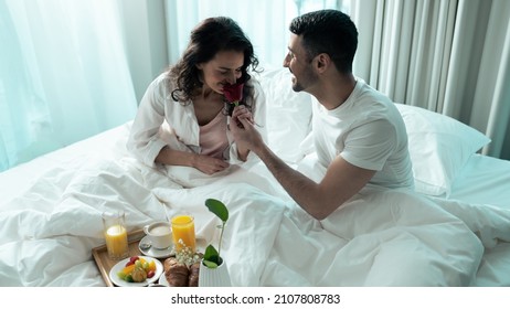 Beautiful Ethnic Couple Sitting on The Bed Wearing White Pyjamas, Eating Breakfast in Bed in a Fancy Hotel Suite Room, Celebrating Valentines Day Together. Female Smelling a Red Rose From Mans Hand. - Powered by Shutterstock