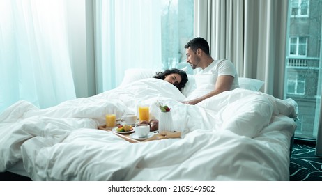 Beautiful Ethnic Couple Spending Valentines Day Together In A Fancy Hotel Suite. Handsome Male Awake, Waking Up His Girlfriend To Surprise Her With The Cute Breakfast Tray Placed On The Bed.