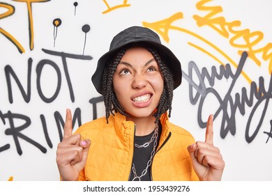 Beautiful Ethnic Hipster Girl With Cheeky Expression Clenches Teeth Points Above Demonstrates Something Against Graffiti Wall Dressed In Fashionable Outfit. Modern Gen Z Teen Has Cool Outlook