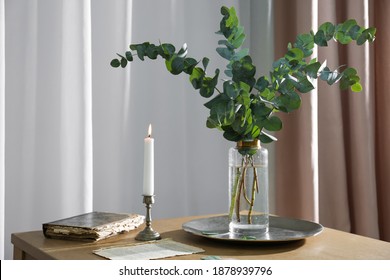 Beautiful eucalyptus branches, old book and holder with burning candle on wooden table indoors. Interior element