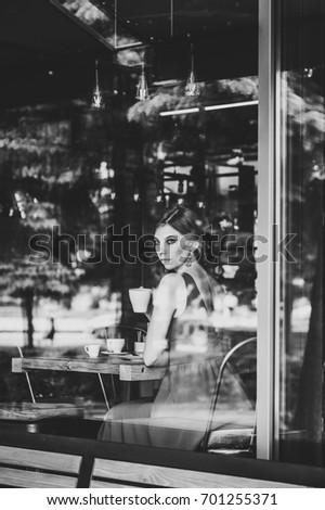 Beautiful European brunette girl in an evening red dress sitting at the window with a cup of coffee at the cafe. Look at the camera through the window. black and white photo
