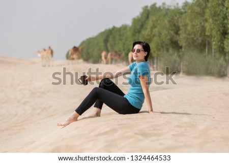 Beautiful European caucasian woman sitting on the sand dune in the desert with sunglasses relaxing drinking a cup of coffee with camels  grazing in the background. 