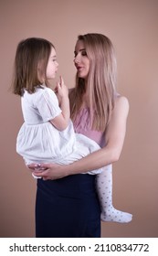 A beautiful European girl in a dress with blonde hair holds and listens to a cute talking girl in a white dress on a brown background in the studio - Shutterstock ID 2110834772