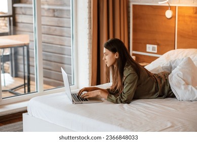 Beautiful european girl lying on bed and using laptop computer. Side view of young smiling woman with brown hair wear pajamas. Concept of rest. Interior of bedroom in modern apartment. Sunny daytime - Powered by Shutterstock