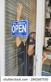 Beautiful European Girl In A Mask Hangs A Sign On A Glass Door Of A Clothing Store (open) After A Cowid-19 Epidemic