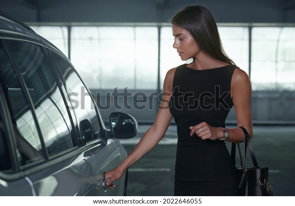 Beautiful\
european girl open door of her private automobile. Serious young\
brunette woman wear black dress and gold handbag. Concept of\
successful modern woman. Inside parking\
lot