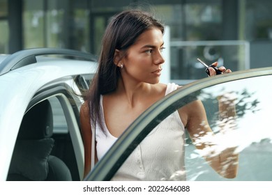 Beautiful european girl stand between door and salon of her private automobile. Serious young brunette woman wear white dress, hold car key and looking somewhere. Concept of modern woman. City daytime