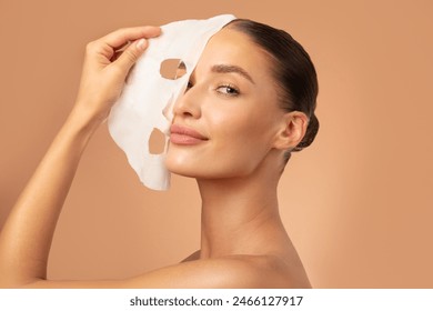 Beautiful European lady removing moisturizing white fabric sheet mask from her face after using it, looking at camera, posing on beige background - Powered by Shutterstock