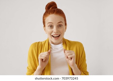 Beautiful European teenage girl with hair knot expressing her astonishment and excitement, clenching fists, raising eyebrows and looking at camera with mouth opened. Cute woman is excited with gift