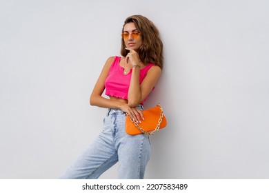 Beautiful European  Woman In Colorful Summer Outfit , Pink Crop Top And Blue Jeans  Posing Over White Background. Holding Orange Bag. Summer Holidays  Fashion Concept.