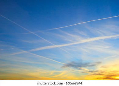 Beautiful evening cloudscape, nature background - Intercrossing airplane contrail against the background of bright blue sky with diffused white clouds in the light of rising sun  
