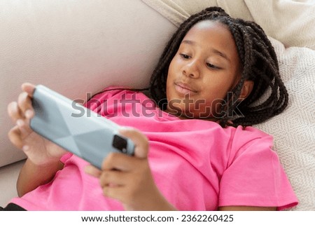 Beautiful, excited African American woman in pink t shirt lying holding mobile phone, playing games, social media, at home in living room. Browsing concept