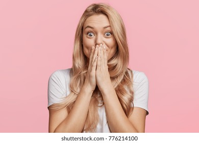 Beautiful excited blonde female covers eyes with happiness, doesn`t expect to recieve pleasant surprise from close friend, being thankful and pleasantly shocked, isolated over pink background