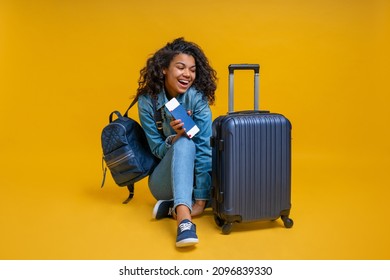 Beautiful excited girl turist sitting on the floor with passport and boarding pass in hands, smiling and looking at her suitcase. Isolated over yellow background. - Shutterstock ID 2096839330