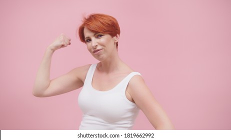 Beautiful excited middle aged redhead woman flexing her muscles, isolated on pink purple background. High quality photo