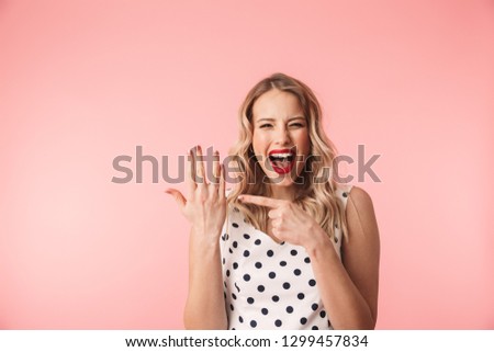 Beautiful excited young blonde woman wearing dress standing isolated over pink background, showing engagement ring on her finger