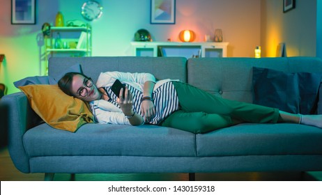 Beautiful Excited Young Girl in Glasses Lying Down on a Couch and Using Her Smartphone. She is Smiling while TV Plays in the Background. Screen Adds Reflections to Her Face Room is Lit with Warm Light - Powered by Shutterstock