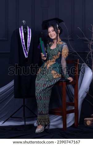 beautiful exotic Asian indonesia woman graduating smiling happily next to her graduation robe wearing a green kebaya and elegant toga strap in an indoor photo studio with a black background