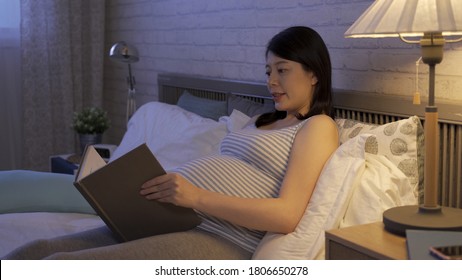 Beautiful Expectant Mom Resting Alone On Bed Is Reading To Her Unborn Baby By Bedside Lamp.