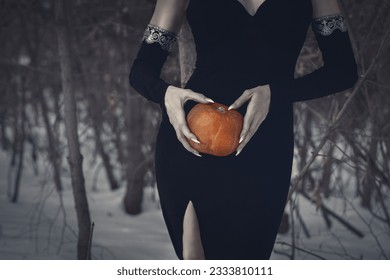 Beautiful faceless woman in black tight dress with a pumpkin in hands in the dark forest, selective focus. Halloween, gothic, witch, November, magic vibes, authentic, athmospheric, dark beauty concept