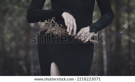 Beautiful faceless woman in black tight dress with a bouquet of dried flowers in hands in the black forest, selective focus. Gothic, vintage, witch, vampire style, autumn mood, dark beauty concept.