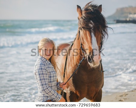 Beautiful fair-haired girl in armor with a horse on the background of the sea. blonde woman near black horse at sea beach. Beautiful woman wearing casual clothing enjoying sunrise morning
