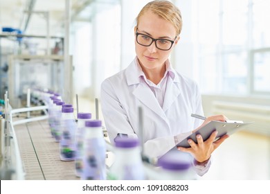 Beautiful Fair-haired Technologist With Clipboard And Pen In Hands Standing At Conveyor Belt And Controlling Production Process At Modern Soy Milk Factory, Portrait Shot