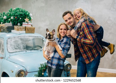 Beautiful Family In Checked Shirts Standing With Dog Near Blue Retro Car With Presents On The Roof. Family In Stylish Decorated Interior Studio For Christmas And New Year Holidays