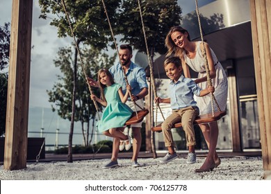 Beautiful family is having fun outside. Parents with children riding on a swing. Mom with dad are playing with their little daughter and son on a terrace with swings.