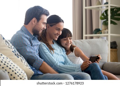 Beautiful Family Of Mom Dad And Little Kid Child Daughter Sitting Together On Sofa Smiling Looking At Smartphone Screen Taking Selfie, Making Video Call Or Recording Vlog With Cellphone At Home