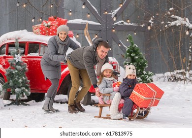 A Beautiful Family (mother, Father, Daughter And Son) Ride A Sleigh, A Snowy Day. On The Background Of A Red Car. Gifts And Christmas Trees.