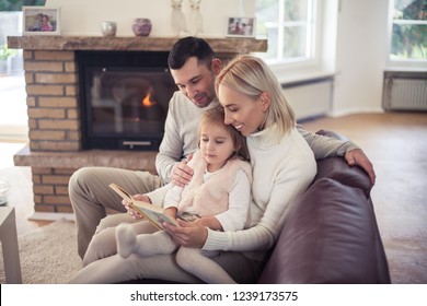 Beautiful Family Sitting By The Fireplace. Mom, Dad And Girl Are Reading A Book. Motherhood. Family. Warm Autumn. Cozy Evening. Home.