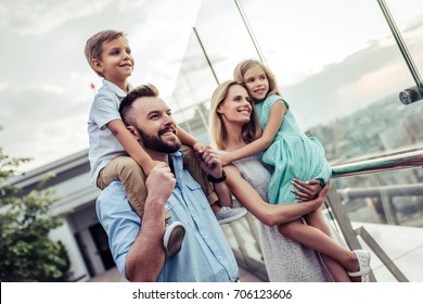 Beautiful Family Is Spending Time Together  Outside. Parents With Their Children On A Roof Terrace With View On A City. Mom And Dad Are Having Fun With Their Charming Daughter And Son.