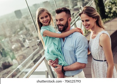 Beautiful Family Is Spending Time Together  Outside. Parents With Their Little Daughter On A Roof Terrace With View On A City. Mom And Dad Are Having Fun With Their Charming Daughter.