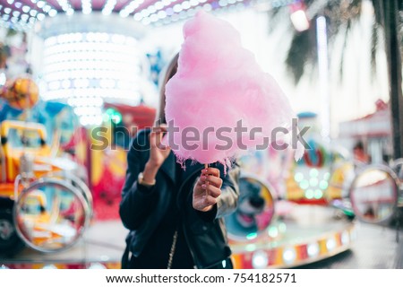 Beautiful fashion model or cute and pretty student woman holds out guilty pleasure, sweet snack, huge pink cotton candy, eats with appetite with fingers, at festival or carnival during summer vacation