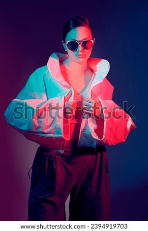 Beautiful fashion model girl posing in stylish white jacket and big sunglasses. Studio shot against a dark background in mixed color light. Bright colors. Fashion and haute couture style.