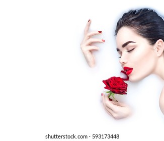 Beautiful Fashion model girl taking milk bath, spa and skin care concept. Beauty young Woman with bright makeup and red rose flower relaxing in milk bath. Healthy Face and hands, red nails and lips