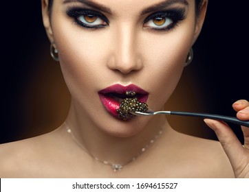 Beautiful Fashion Model woman eating black caviar. Beauty girl portrait with caviar on her lips. Fashion female with spoon of black Caviar isolated on Black background, closeup