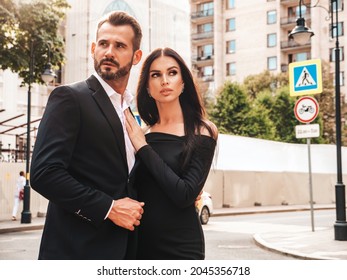 Beautiful Fashion Woman And Her Handsome Elegant Boyfriend In Suit. Sexy Brunette Model In Black Evening Dress. Fashionable Couple Posing In The Street In Europe. Brutal Man And His Female Outdoors