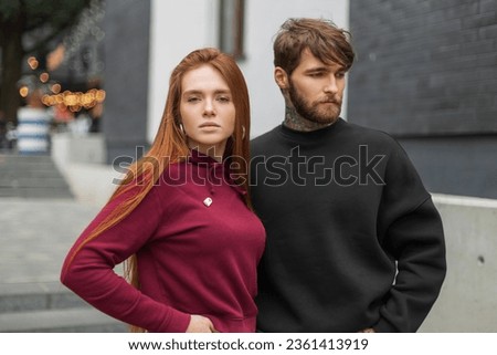 Beautiful fashion young stylish couple in trendy street outfit with hoodies on the street. Pretty red-haired fresh girl and a handsome brutal bearded man in a black sweatshirt